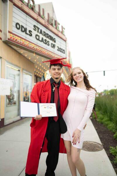 A graduate in a red cap and gown stands smiling with his diploma beside a woman in a light dress.